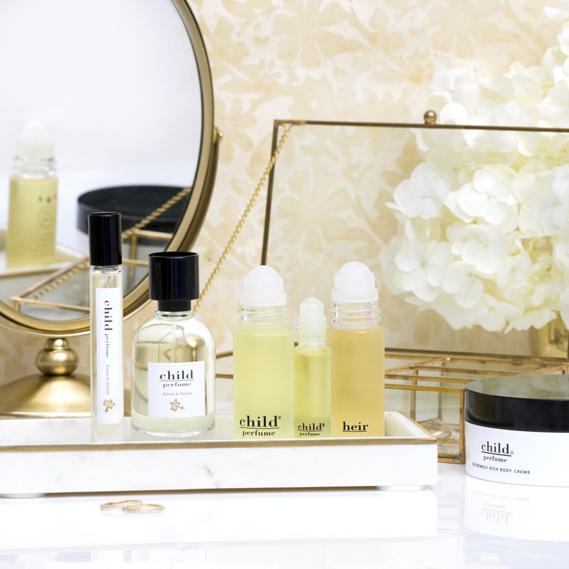 Lifestyle shot of a selection of Child Perfume products including Roll Ons, Limited Edition Spray, Travel Spray and Body Creme on vanity tray with white flowers and mirror in the background