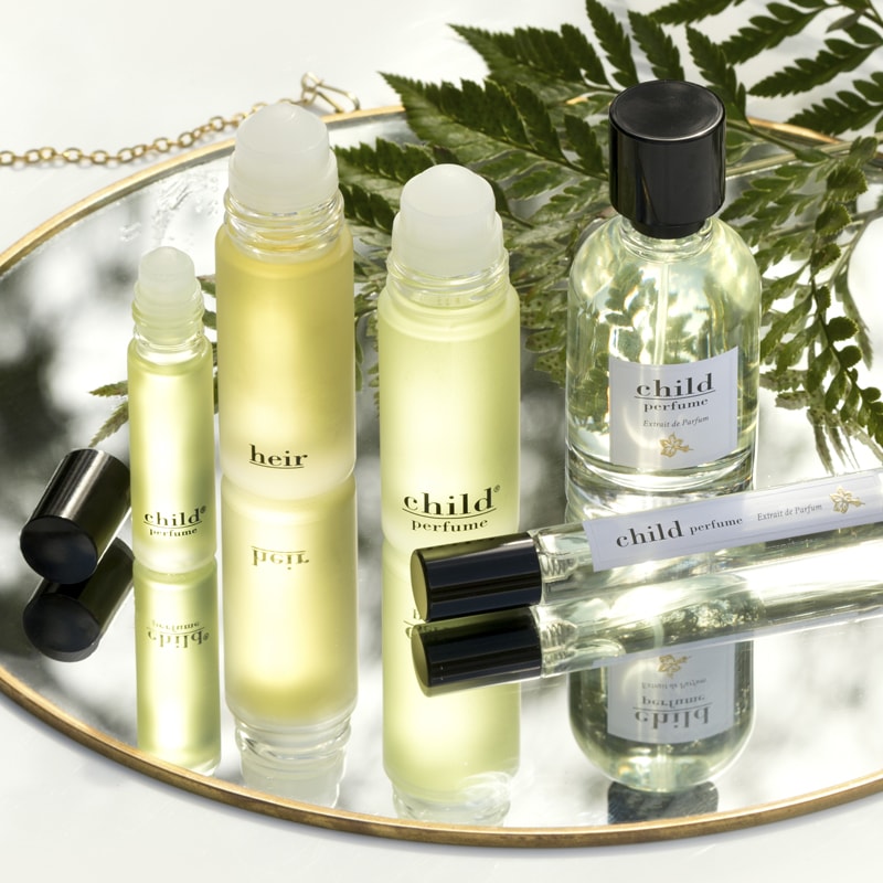 Lifestyle shot of a selection of Child Perfume products including Roll Ons, Limited Edition Spray and Travel Spray on mirrored vanity tray and fern branch in the background